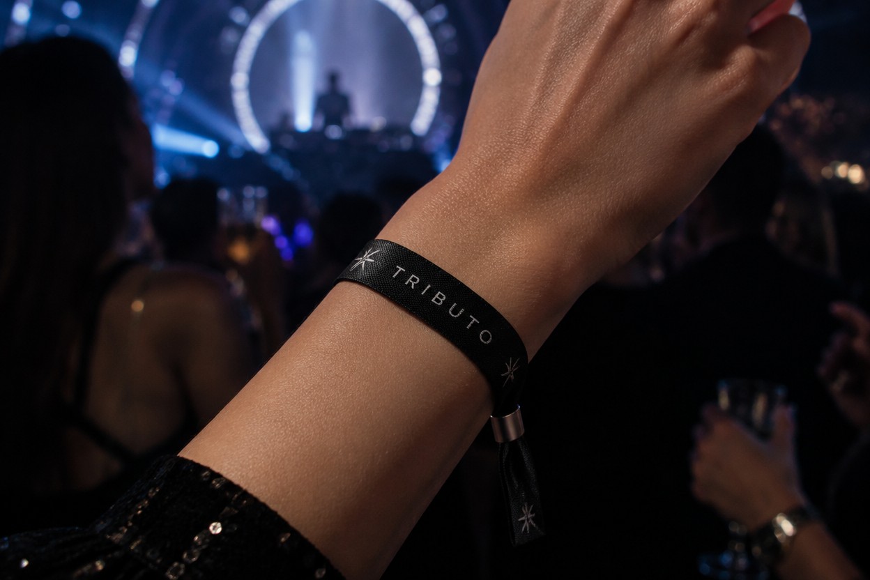 Premium black fabric event wristband worn on a person's wrist at an elegant electronic music event. Minimal black and titanium design, subtle metallic details, sophisticated nightclub environment, dark cinematic lighting, electric blue and magenta reflections, realistic fabric texture, high-end nightlife photography, contemporary and luxurious aesthetic. Apply the uploaded TRIBUTO wristband artwork exactly as provided, preserving the logo, typography, proportions and colors. Do not alter the artwork.